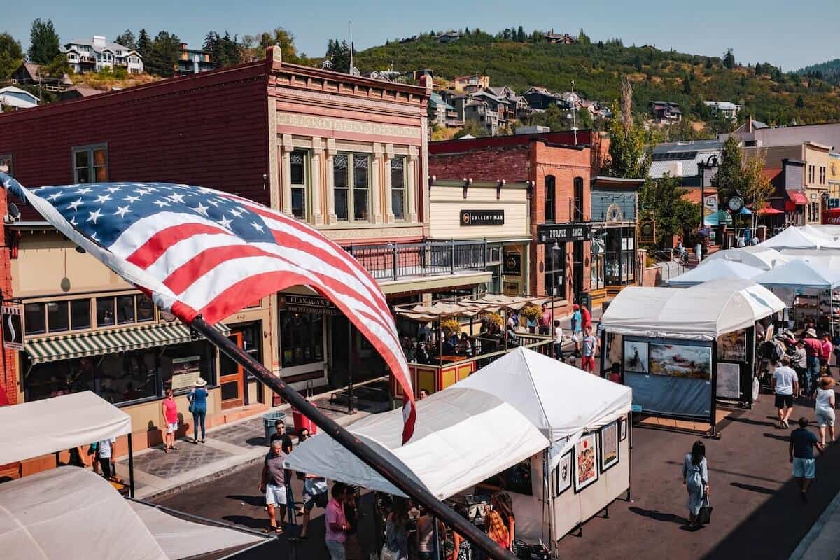 Cover photo from Things to do in Park City Utah featuring an aerial view of main street in Park City in summer with an American flag in the foreground and white tents of the market lining the street