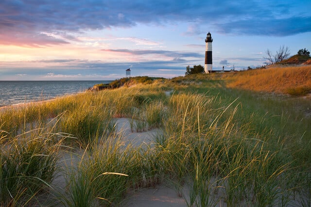 Grassy sand in the foreground with the black and while ringed Sable Point Lighthouse in the distance with the ocean behind that