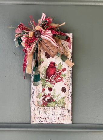 Winter cardinal napkin decoupaged to a distressed white background with a red and green messy bow and a hangtag that says "Believe". Cardinal Napkin Sign