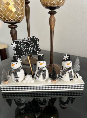 Snowmen Ornament Shelf sitter made with 3 mini felt white snowmen from walmart, on a wood base with a chalkboard sign that says "let it snow", snowballs made with white pom poms, and black and silver bottle brush christmas trees.