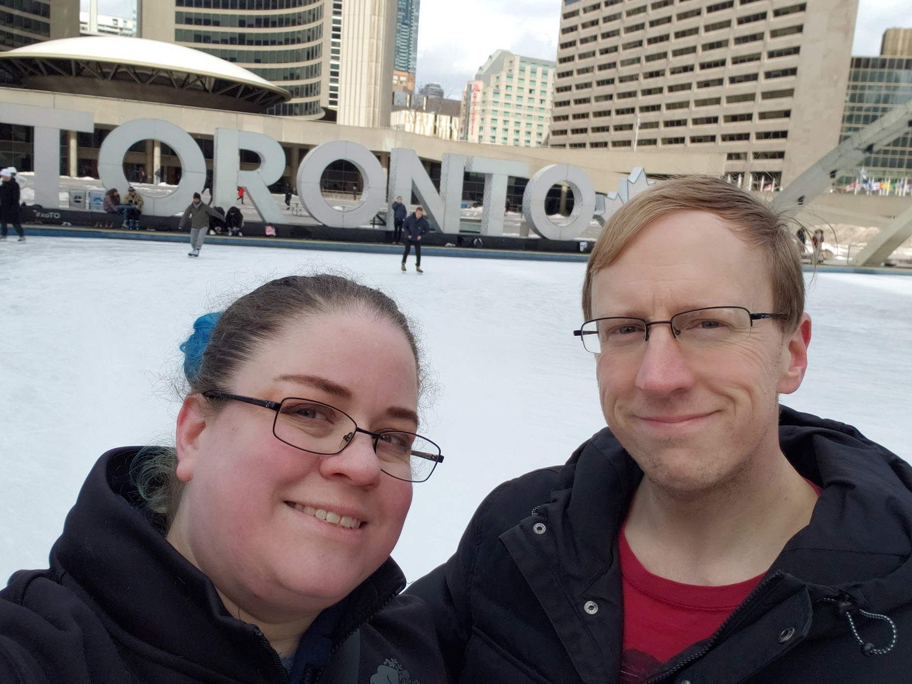 A middle aged couple taking a selfie in front of the ice rink at Nathan Phillips Square.