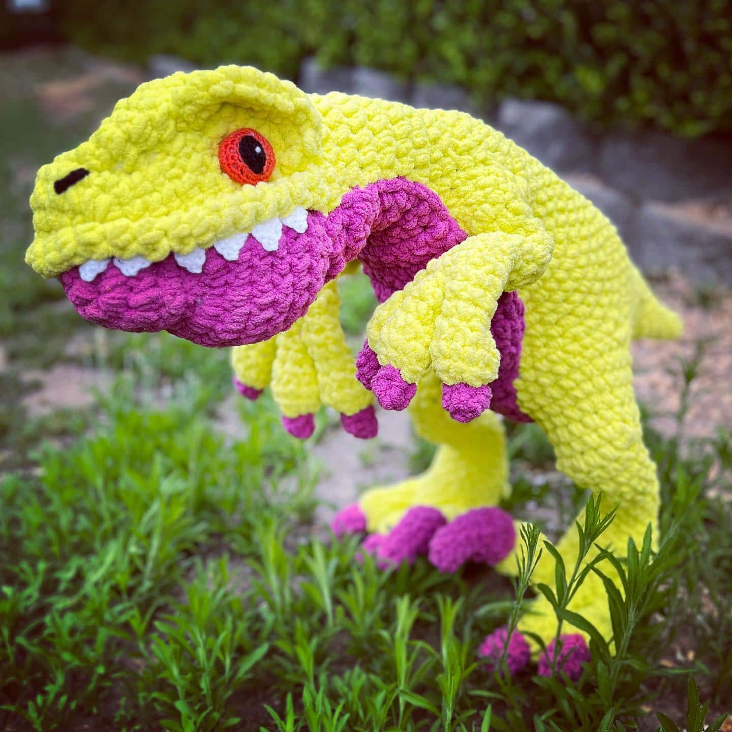 Rori the Raptor, a crocheted dinosaur plush in bright yellow and purple, stands on grass. With an orange eye and playful expression, its mouth is slightly open to reveal white teeth. The background features greenery and stone paving, creating a charming setting for this delightful crochet pattern design. -Marly Bird