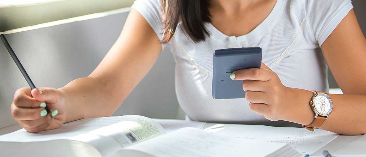 A person writing in a workbook while holding a calculator.