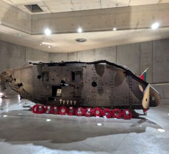 Rusty brown wreckage of a WW1 tank, set in a concrete display hall. A row of red wreaths are laid in front of it.