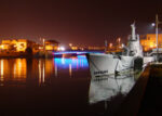 Submarine moored alongside in still water at night with nearby coloured street lights reflecting on the water