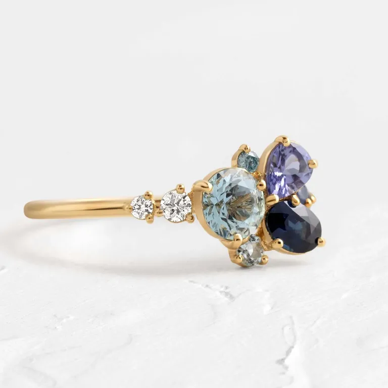 Clear water cluster Taylor Swift inspired engagement ring with aquamarine, sapphire, and tanzanite, accented by white diamond, aqua blue diamond, aquamarine, and sapphire side stones from Melanie Casey