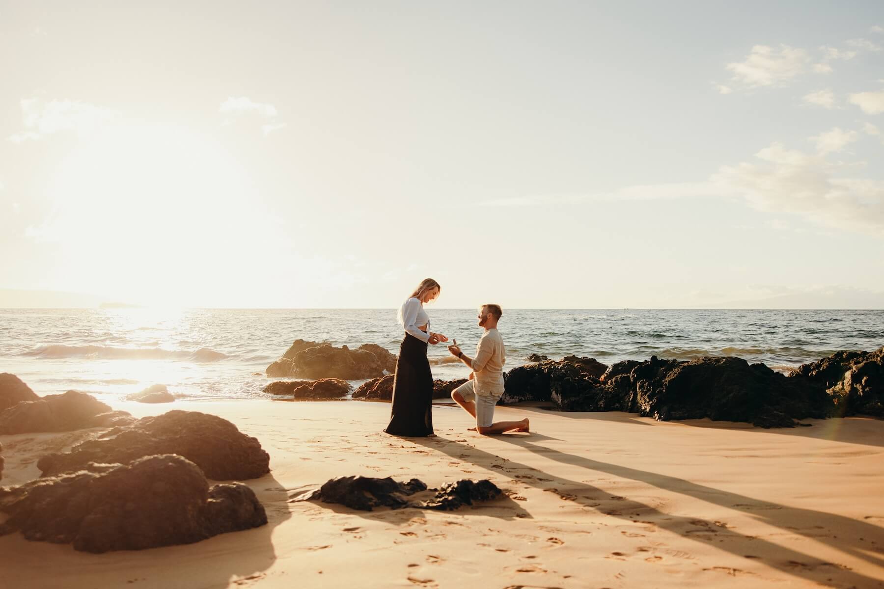 A man is on one knee proposing to his girlfriend on the beach in Maui while the sun sets.