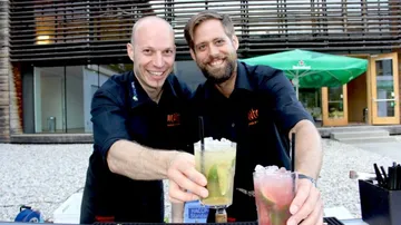 Zwei Bartender präsentieren Cocktails vor einem modernen Gebäude.