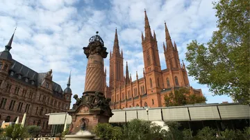 Säule steht vor rotem Gebäude, umgeben von Bäumen und Wolken.