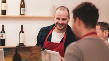 Auf dem Bild sind drei junge Männer zu sehen, welche auf ein, von ihnen gemaltes Bild, schauen. Im Hintergrund sind Weinflaschen auf Holzregalen zu sehen