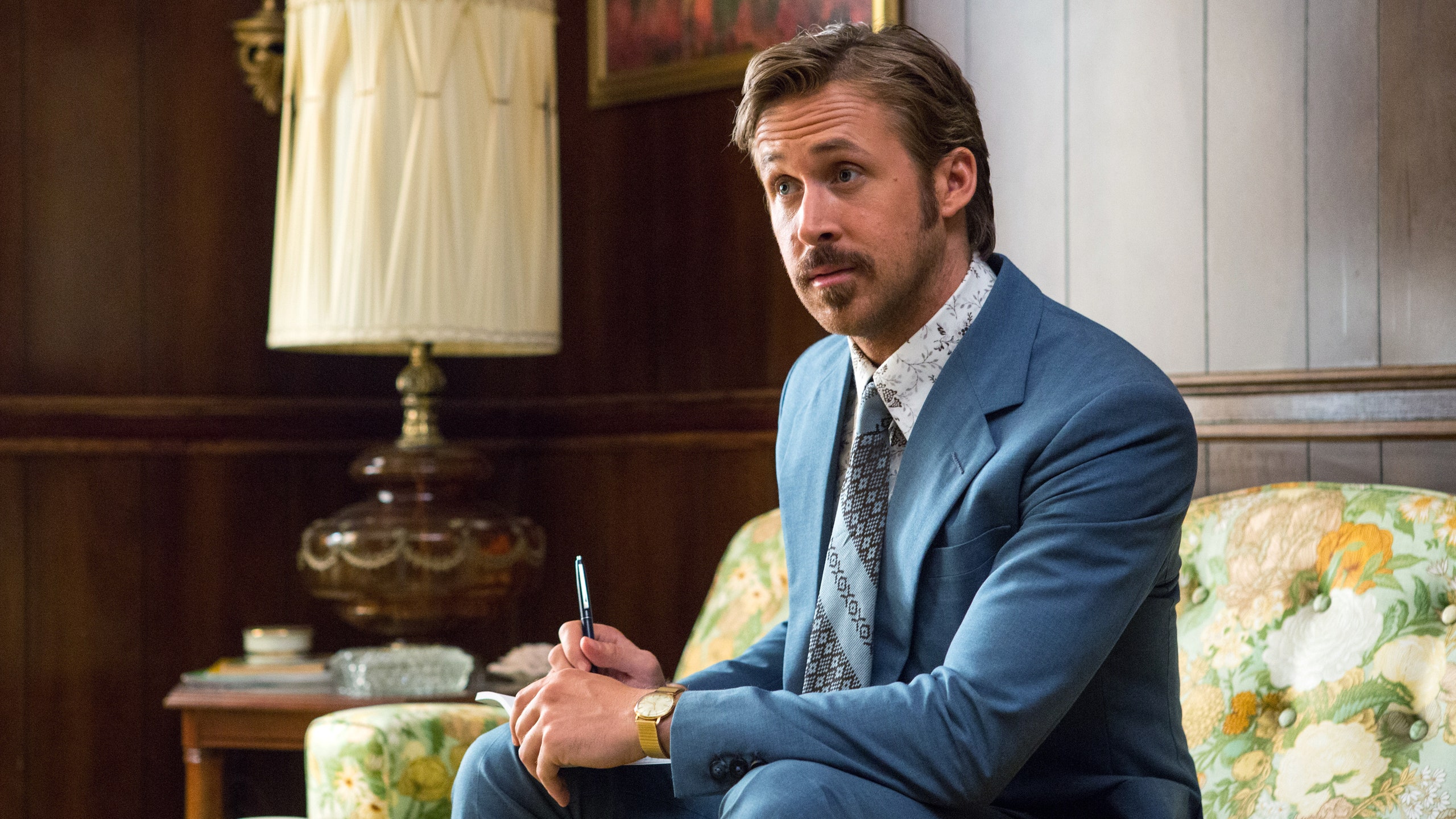 This image may contain Ryan Gosling Human Person Coat Suit Clothing Overcoat Apparel Lamp Table Lamp and Sitting