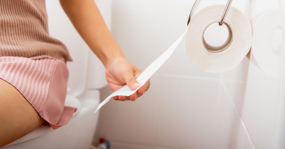 A woman wearing a pink dress sits on a toilet and grabs for some toilet paper. 