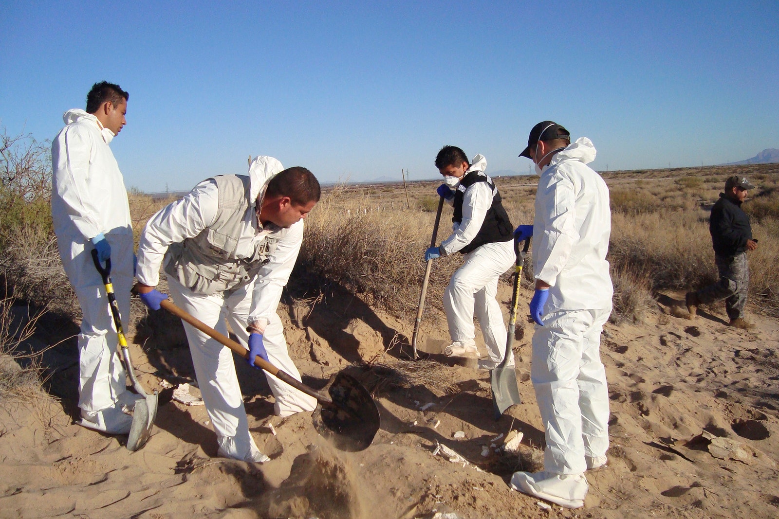 People in white hazmat suits digging in the desert with shovels.