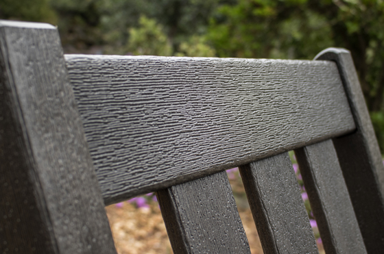 Close-up of the top part of a vintage plank-style tabletop vintage bench, illustrating its textured surface and dark finish. The background is blurred, suggesting a natural setting with greenery and some purple flowers-a perfect addition to a modern home.