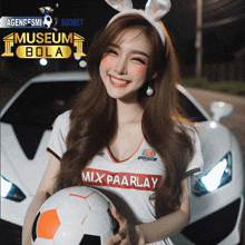 a woman wearing a mix paarlay jersey holds a soccer ball in front of a car