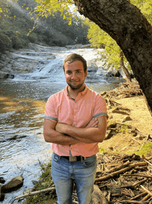 a man in a pink shirt stands in front of a waterfall with his arms crossed