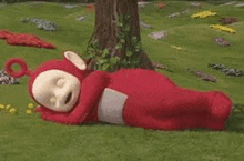 a red teletubbies teddy bear is laying down in the grass .