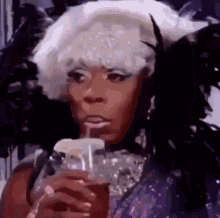 a drag queen is drinking a drink through a straw while wearing a feathered headpiece .