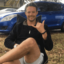 a man sitting in front of a blue car giving the thumbs up sign