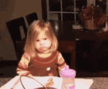 a little girl is sitting at a table with a bowl of food and a purple bottle .