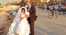 a man in a suit stands next to a little girl in a wedding dress holding a bouquet of flowers