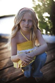 a young girl wearing a yellow top and blue jeans sits on a wooden dock