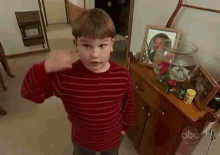 a young boy in a red striped shirt is standing in front of a dresser with a picture on it .