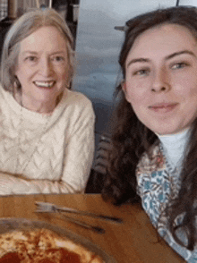 two women are posing for a picture while sitting at a table with a pepperoni pizza .