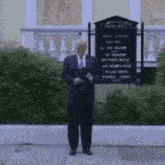 a man in a suit and tie is standing on a sidewalk in front of a sign that says " all are welcome "