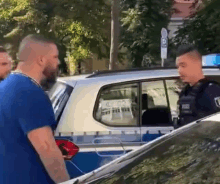 a man in a blue shirt is standing in front of a police car talking to a police officer .