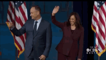 a man in a suit and tie and a woman in a purple suit are on a stage in front of american flags