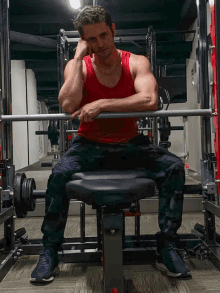 a man in a red tank top sits on a bench holding a barbell in a gym