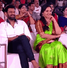 a man and a woman are sitting in front of a crowd . the woman is wearing a green saree .