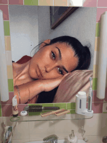 a woman laying on a pillow in front of a mirror with a bottle of toothpaste on the shelf