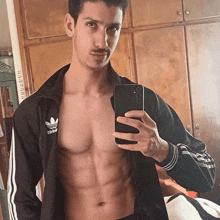 a shirtless man is taking a selfie with his phone while wearing a black adidas jacket