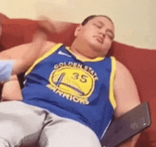 a man wearing a golden state warriors jersey is laying on a couch with a laptop .