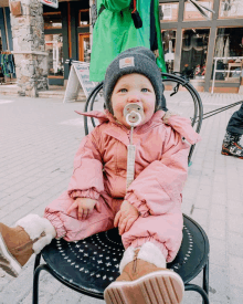 a baby wearing a pink snowsuit and a pacifier sits on a chair