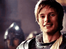 a young man wearing armor is smiling with a helmet in the background