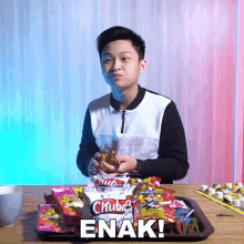 a young man is sitting at a table with a tray of snacks and the words " enak " on the bottom