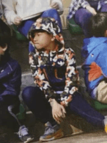 a young boy wearing a camouflage hoodie is sitting on the ground in a crowd of people .