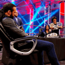 a man sits in a chair in front of a table with a woman holding a wwe championship behind him