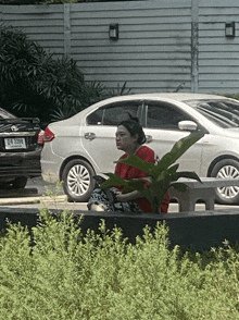 a woman in a red shirt sits in front of a white car with a license plate that says jm 9298