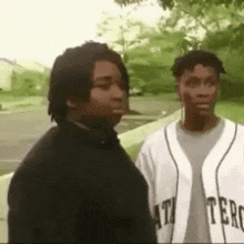 two men are standing next to each other on a sidewalk . one of the men is wearing a baseball jersey .