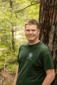 a young man in a green t-shirt is standing next to a tree in the woods .