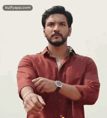 a man with a beard wearing a red shirt and a watch is standing in front of a white wall .