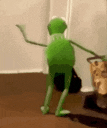 a green kermit the frog is dancing on a wooden floor in a room .