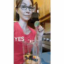 a woman in a red shirt is holding a piece of fruit in front of a blender that says yes