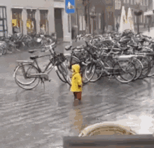 a little girl in a yellow raincoat is standing in the rain in front of a row of bicycles .