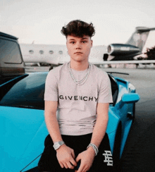 a young man wearing a givenchy t-shirt is sitting in front of a blue car