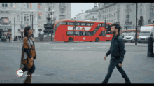 a man and a woman are walking in front of a red double decker bus with a zeus logo on the bottom